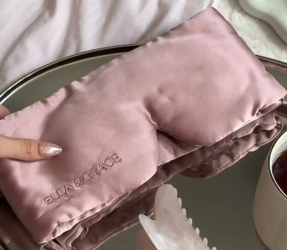 A pink silk sleep mask with a plush lining and the brand name 'ELLA & GRACE' embroidered on the front, held by a hand over a silver tray and other spa accessories.