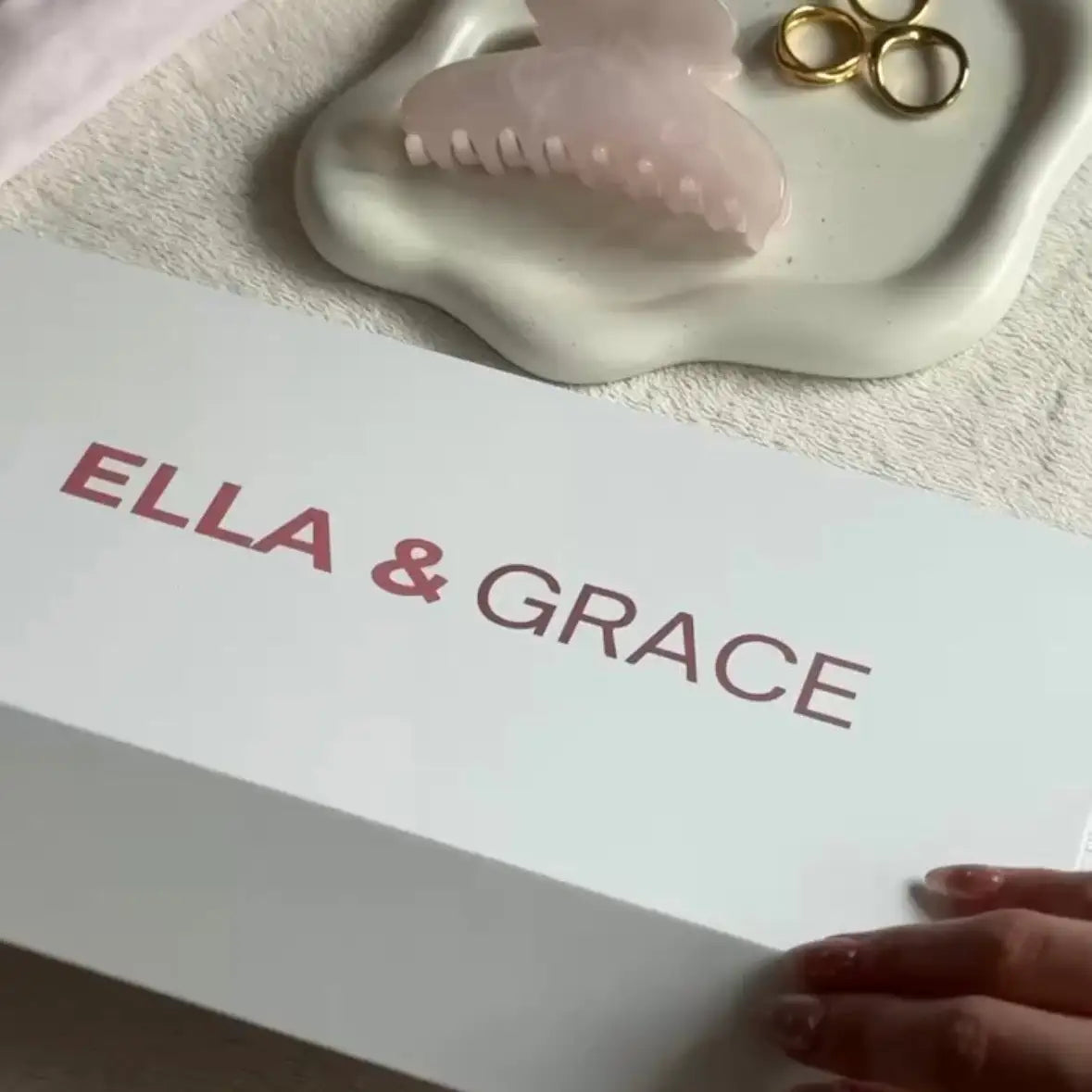 A hand holds a white box printed with the brand name 'ELLA & GRACE' in pink. In the background, a pale pink claw hair clip rests on a white dish with two gold rings, all on a soft beige fabric surface.
