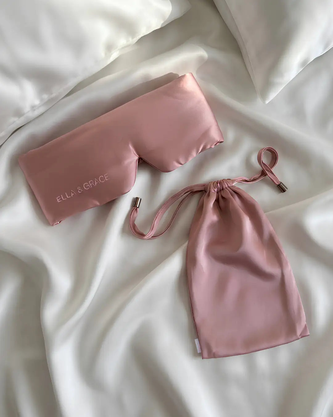 A pink silk sleep mask and matching drawstring pouch laid out on white silk bedding. The sleep mask has the brand name 'ELLA & GRACE' embroidered on it.